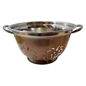 May include: A stainless steel and copper-toned colander with two handles. The copper-toned sides feature decorative cut-out designs. The colander is designed for draining liquids from food items.