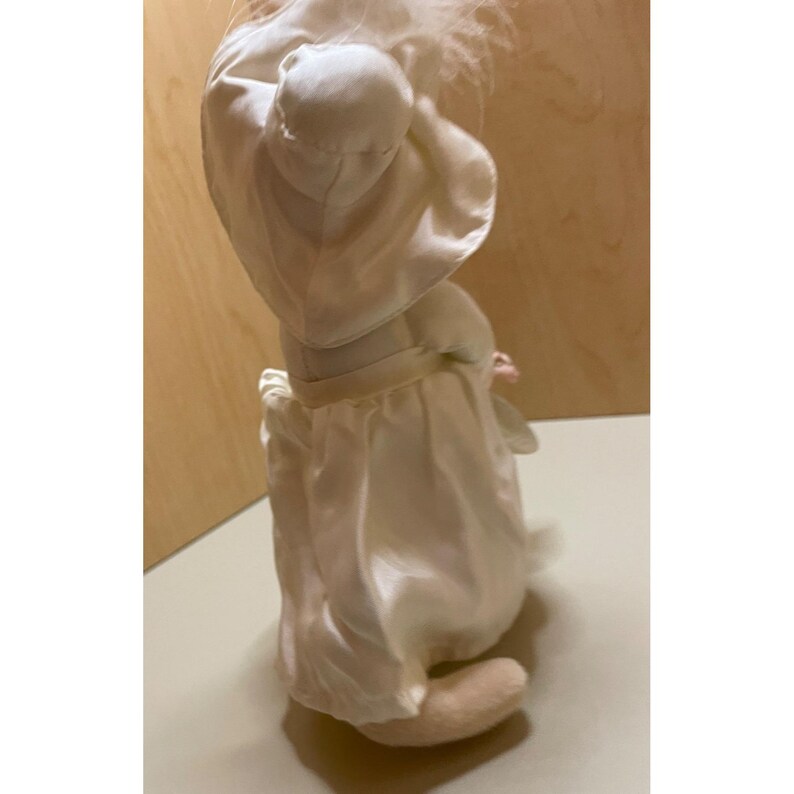 May include: A white porcelain doll dressed in a white satin gown with a white feathered halo. The doll is holding a white satin flower in its hand.