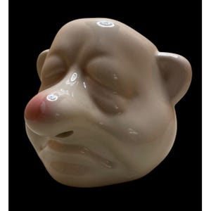 May include: A white ceramic sculpture of a face with a large, pink nose and closed eyes. The face has a sad expression with a downturned mouth.
