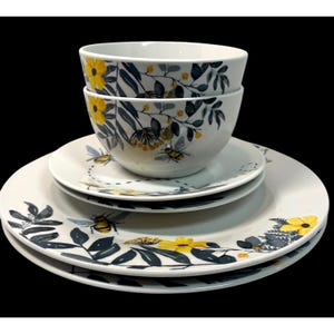 May include: A white ceramic dinnerware set featuring a floral and bee design. The set includes two bowls and three plates of varying sizes. The design incorporates yellow flowers, blue leaves, and stylized bees. The dinnerware is stacked on a black surface.
