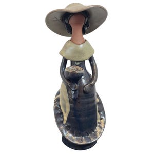 May include: A ceramic figurine of a woman wearing a wide-brimmed hat and a long dress. The figure is primarily dark blue with tan and brown accents. The woman is holding a bouquet of flowers. The figurine is a decorative item.