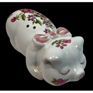 May include: White ceramic pig figurine with pink bow and floral accents. The pig has a whimsical design with painted pink and purple flowers and green leaves. The figurine has two holes on its back.