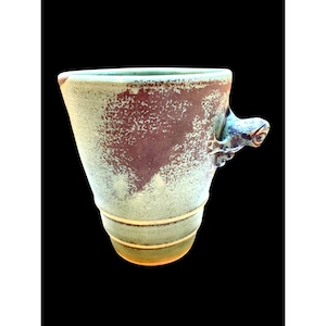 May include: Handmade ceramic mug with a unique design. The mug features a light green and brown speckled glaze, with a frog-shaped handle. The mug has a flared shape and is approximately 5 inches tall.