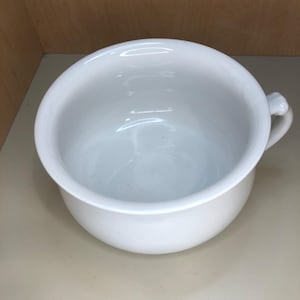 May include: A white ceramic chamber pot with a rounded body and a small handle. The interior is smooth and the exterior is glossy. The chamber pot is sitting on a light-colored surface.
