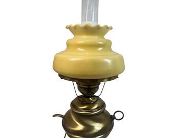 May include: A table lamp featuring a brass base with a rounded, metallic body, a small side handle, and a ring attachment. The lamp is topped with a pale yellow, ruffled glass shade and a clear, cylindrical glass chimney insert.