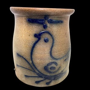 May include: A tan stoneware pottery jar with a dark blue bird design. The jar has a cylindrical shape with a slightly flared rim. The bird is stylized, with a curved body and a small dot for an eye. The design is hand-painted.