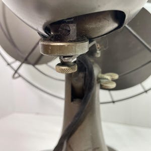 May include: Close-up of an antique metal fan. The fan has a dark gray finish with a wire cage and a textured knob. The fan's design is vintage, with visible wear and tear, suggesting age and use.