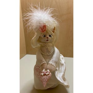 May include: A white porcelain cat figurine wearing a white hat with a pink rose and white feathers. The cat is holding a pink heart-shaped sign that says "Cat-N-Us".