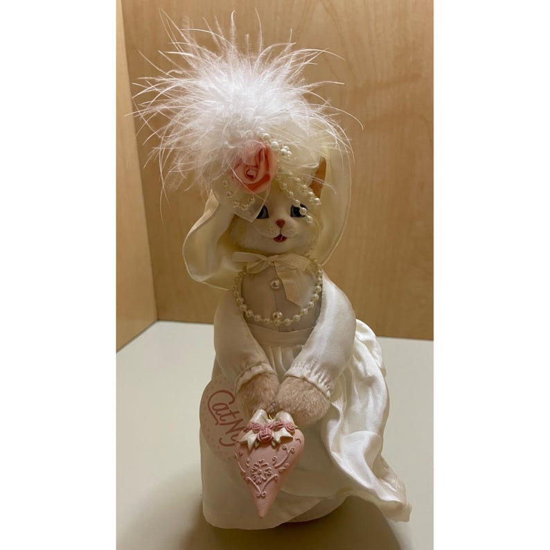 May include: A white porcelain cat figurine wearing a white hat with a pink rose and white feathers. The cat is holding a pink heart-shaped sign that says "Cat-N-Us".