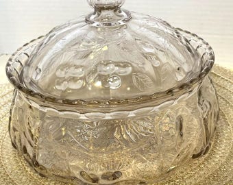 May include: A clear glass candy dish with a lid. The dish and lid are decorated with a cherry and leaf pattern. The lid has a small, round knob on top for easy handling. The dish is round with a scalloped edge.