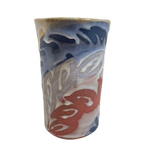 May include: A handmade ceramic tumbler with a unique design. The cup features a blend of colors, including blue, red, and white, with a textured, abstract pattern. The rim has a light brown color.