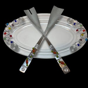 May include: A clear oval serving platter with two crossed salad servers. The servers have silver handles with colorful beads and wire detailing. The platter's rim is decorated with silver wire swirls and colorful beads. The background is black.
