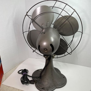 May include: A vintage, gray metal electric fan with four blades, a protective wire cage, and a rounded base. The fan has a dark power cord and plug. The fan's design is reminiscent of mid-century appliances.