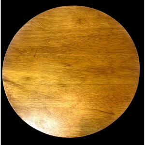 May include: A circular wooden board featuring a smooth, polished surface with visible natural grain patterns in shades of light to medium brown. The wood displays subtle variations in color and texture across its flat, round surface.
