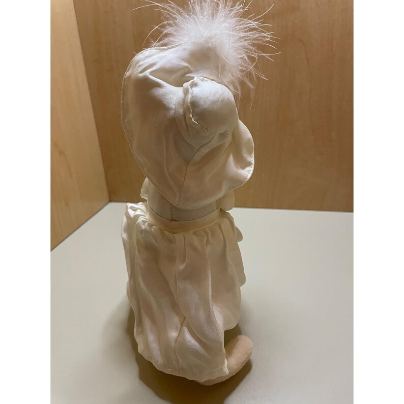 May include: A white satin doll with a white feather hat. The doll is wearing a white satin dress with a white satin sash.