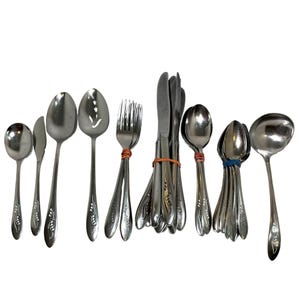 May include: A collection of stainless steel silverware, including spoons, forks, knives, and a ladle. The silverware has a floral design on the handles and is bundled together with rubber bands. The utensils are arranged on a white background.