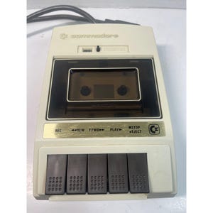 May include: A vintage Commodore cassette player in off-white with a black cassette tape compartment. The player has black buttons for record, rewind, fast forward, play, stop, and eject. The Commodore logo is visible on the top.
