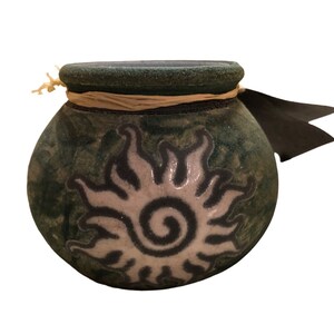 May include: A green ceramic pot with a white sun design and a brown twine accent. The pot has a black ribbon attached to the side.