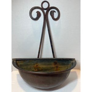 May include: A wall-mounted metal planter featuring a semi-circular basin with a dark, weathered patina and visible rust. The back plate consists of two long, curved metal rods that meet at a central ring, creating a decorative scrollwork design.