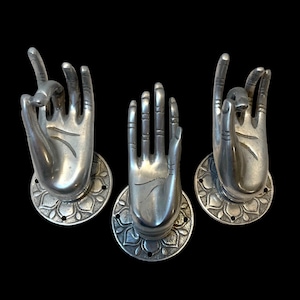May include: Three silver-colored hand sculptures in various mudra positions. Each hand is mounted on a circular base with a floral design. The background is solid black, highlighting the metallic finish of the sculptures.
