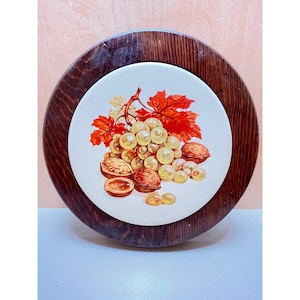 May include: Round wooden trivet with a white ceramic center. The ceramic features a still life of grapes, walnuts, and autumn leaves. The trivet has a dark brown wooden frame. A decorative kitchen item.