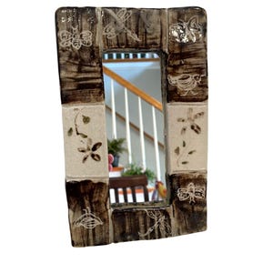 May include: Rectangular ceramic mirror with a brown and cream-colored frame. The frame is decorated with floral and insect motifs. The mirror reflects a glimpse of a staircase and interior. The frame is approximately 6 inches wide and 8 inches tall.