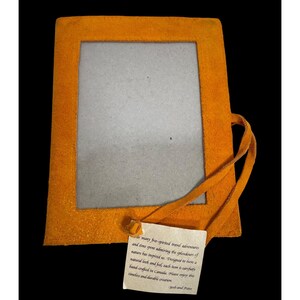May include: An orange suede photo frame with a rectangular opening. A leather tie is attached to the side. A small tag with text is attached to the frame. The frame is designed to have a natural look and feel.