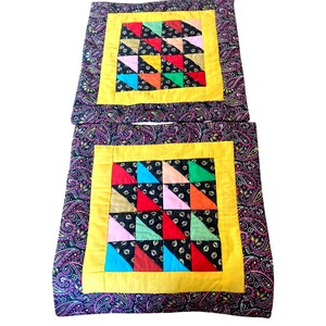 May include: Two square quilted cushion covers featuring a central patchwork design of multicolored triangles in red, blue, green, pink, and gold on a black background. A solid yellow border surrounds the center, framed by a dark paisley print.