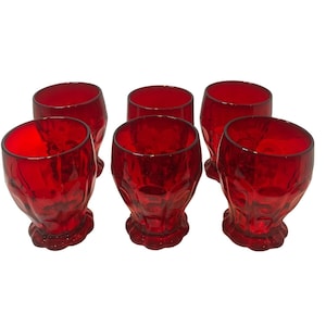 May include: Six red-colored drinking glasses. Each glass has a rounded bowl shape with a textured, patterned design and a short, decorative stem. The glasses are made of a transparent material, allowing light to pass through.