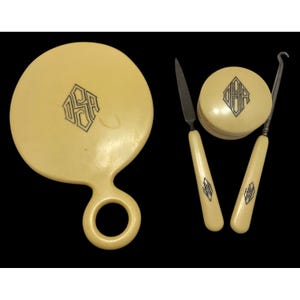 May include: An antique vanity set in a cream color, featuring a round hand mirror, a small round container, and two tools. Each piece has a black monogram design. The set is displayed against a black background.