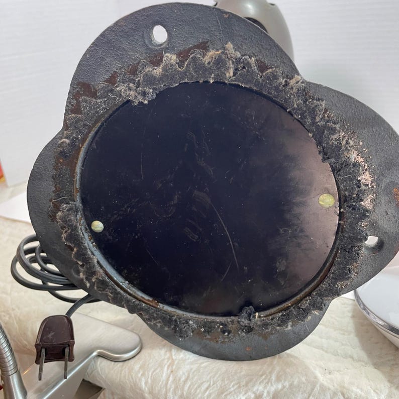 May include: Underside view of a vintage black metal lamp base with a circular center and a decorative, scalloped edge. The base has a three-prong plug and a black cord. The metal shows signs of age and wear.