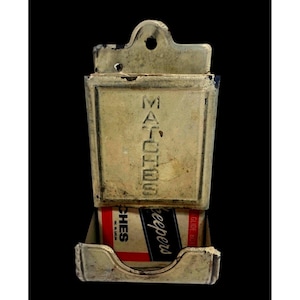 May include: A vintage wall-mounted metal match holder with a distressed cream finish. The front panel is embossed with the word MATCHES in vertical capital letters. A box of Match Keepers matches is visible in the lower tray, featuring red and black text.