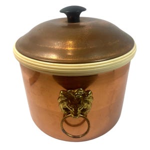 May include: A copper ice bucket with a lid and a black knob. The bucket has a cream-colored rim and a gold lion's head with a ring handle. The copper has a shiny finish.