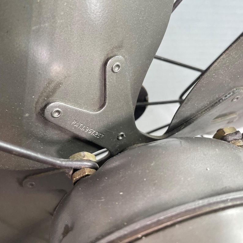 May include: Close-up of a vintage gray electric fan. The fan blades and housing are a muted gray color. The metal frame has the text "PAT.1967257" stamped on it. The fan blades are protected by a wire cage.