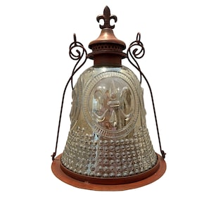 May include: A clear glass lantern with a fleur-de-lis design, surrounded by a metal frame and a copper-colored top. The lantern has a bell shape with a round base and decorative accents.