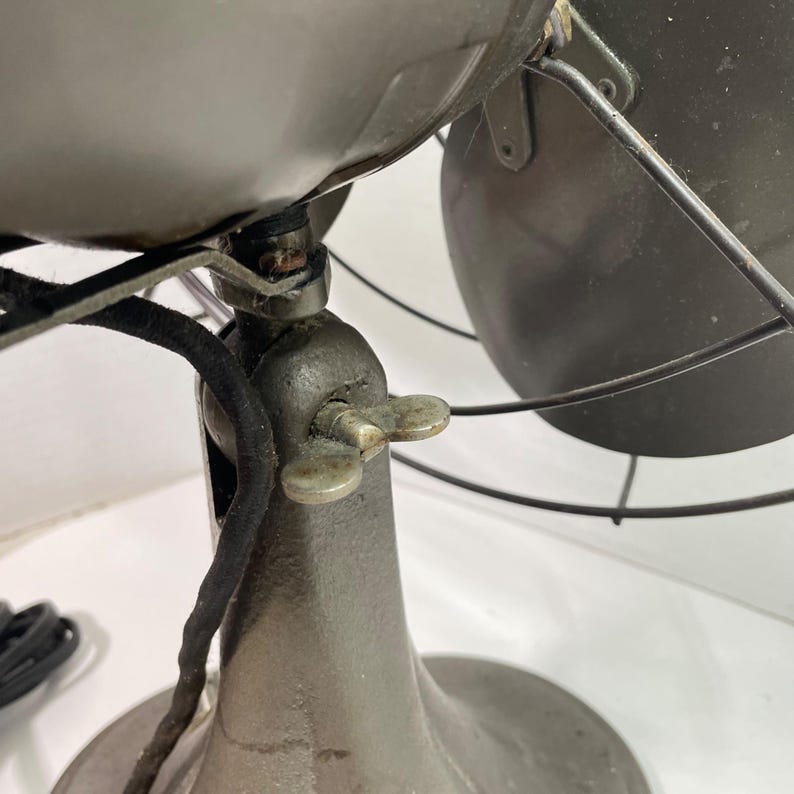 May include: Close-up of an antique gray metal fan. The fan has a rounded base, a central support with a wingnut, and a protective cage around the blades. A black cord extends from the fan's motor.