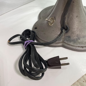 May include: Close-up of an antique metal lamp base with a black power cord and a brown plug. The base is a dark gray color with a decorative, scalloped edge. A purple rubber band secures the cord.