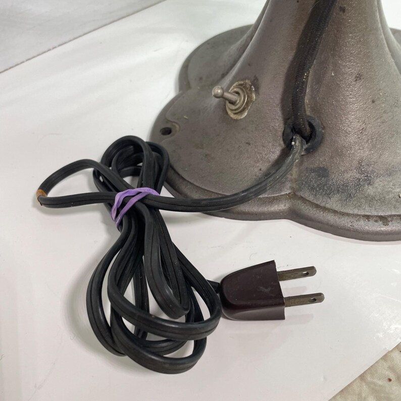May include: Close-up of an antique metal lamp base with a black power cord and a brown plug. The base is a dark gray color with a decorative, scalloped edge. A purple rubber band secures the cord.