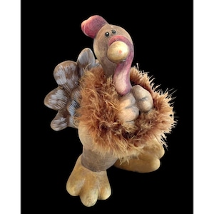 May include: A whimsical ceramic turkey figurine with a brown body, tan feet, and a fluffy brown feather collar. The turkey has a red wattle and comb, and a tan beak. The turkey is a decorative item.