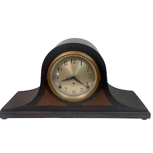 May include: A dark wood mantel clock with a gold-tone clock face. The clock has a curved top and a rectangular base. The clock face features black numerals and hands. The clock is approximately 12 inches wide.