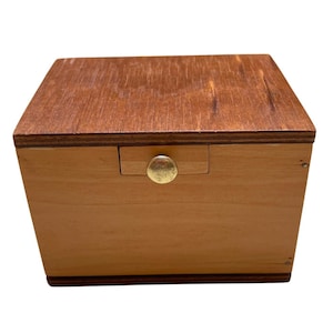May include: A small wooden box with a drawer and a brass knob. The box is rectangular with a light brown body and a darker brown top. The drawer is centered on the front of the box.