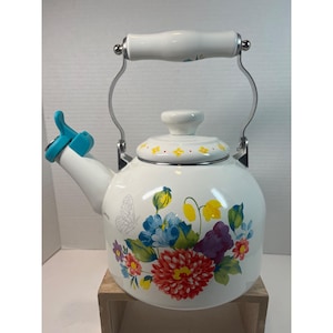 May include: White enamel tea kettle with a floral design featuring red, blue, and yellow flowers, and a butterfly. The kettle has a white handle and a blue whistle. The handle is 7 inches long.