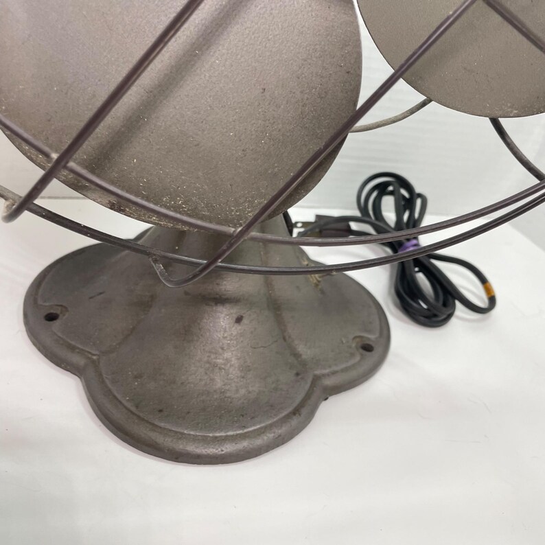 May include: An antique, gray metal fan with two blades, encased in a wire cage. The fan has a decorative base and a black power cord. The fan is designed for air circulation.