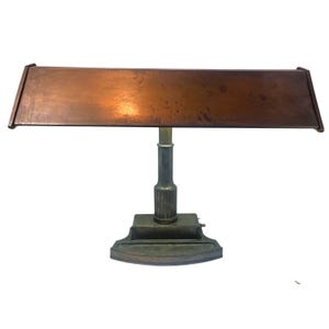May include: A vintage desk lamp with a copper shade and a brass-colored base. The rectangular shade is angled downward, and the lamp has a classic, industrial design. The base is rectangular with a decorative pattern.