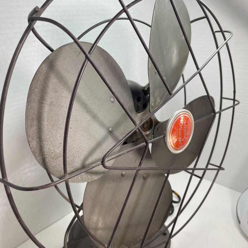 May include: Vintage Emerson electric fan with a metal cage and blades. The fan has a dark gray finish with a red and white Emerson label. The blades are silver and the cage is a dark gray color.
