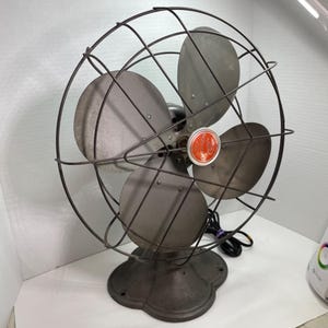 May include: Vintage Emerson electric fan with three blades and a protective wire cage. The fan has a dark gray finish and a round base. A red and white Emerson label is visible on the center of the fan.