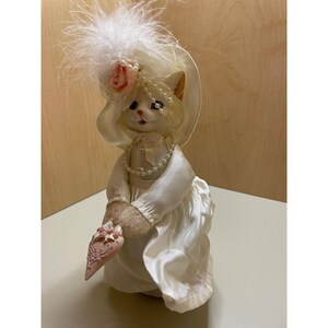 May include: A white cat doll wearing a white dress and hat with a pink rose. The doll is holding a pink heart-shaped object with a floral design.