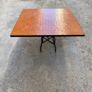 May include: Square orange mosaic tile table with black metal legs. The tabletop has a glossy finish and a decorative pattern. The table is approximately 30 inches square and is suitable for indoor or outdoor use.