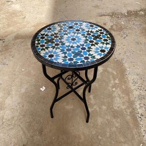 Może przedstawiać: Okrągły stół z mozaiką i czarną metalową ramą. Blat ma geometryczny wzór w odcieniach niebieskiego, białego i turkusowego. Stół ma średnicę około 61 cm i stoi na czterech zakrzywionych nogach.