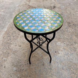 Może przedstawiać: Okrągły stół z mozaiki z czarną podstawą z kutego żelaza. Blat ma geometryczny wzór z niebieskich, zielonych i białych płytek. Stół ma średnicę około 61 cm i stoi na czterech zakrzywionych nogach.